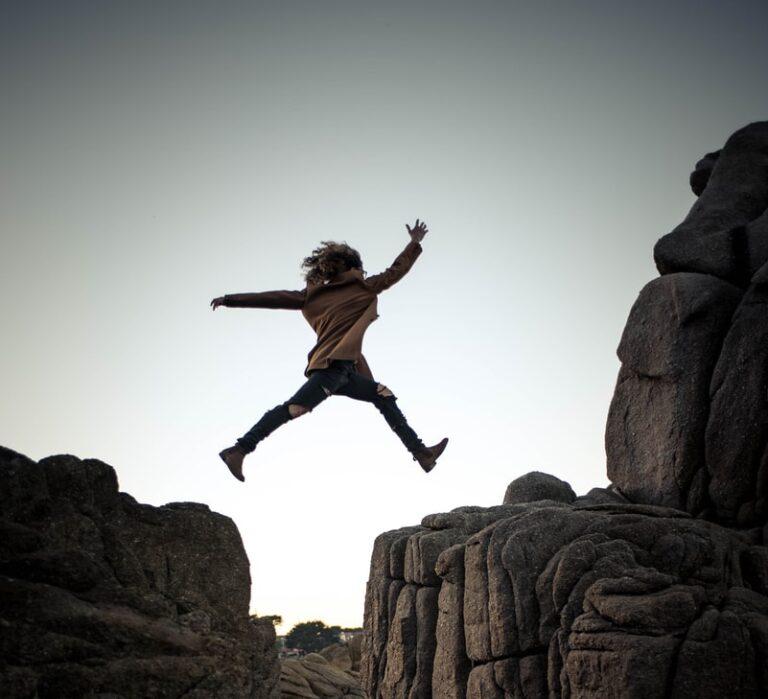 person jumping on big rock under gray and white sky during daytime